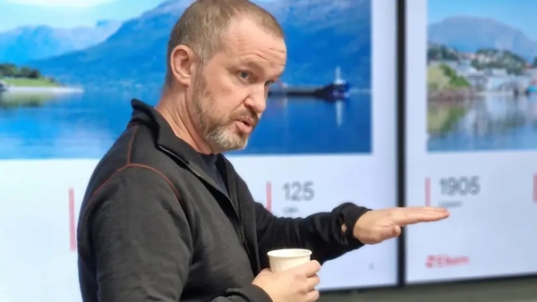 A person stands in front of a presentation screen displaying images of Norwegian landscapes, holding a cup and gesturing with one hand.