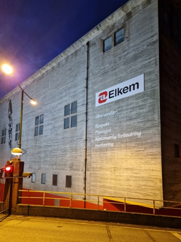 A factory building at dusk with an Elkem sign mounted on the wall, illuminated by streetlights.