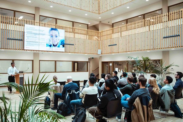 Group of people seated in a large room looking at a screen as someone on stage gives a presentation 