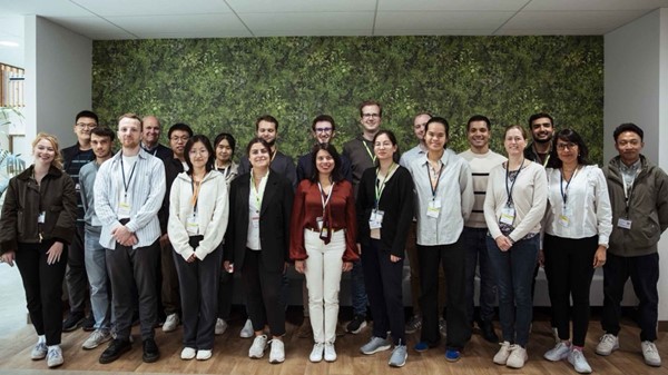 Large group of young people standing in an office setting in front of a green patterned wall