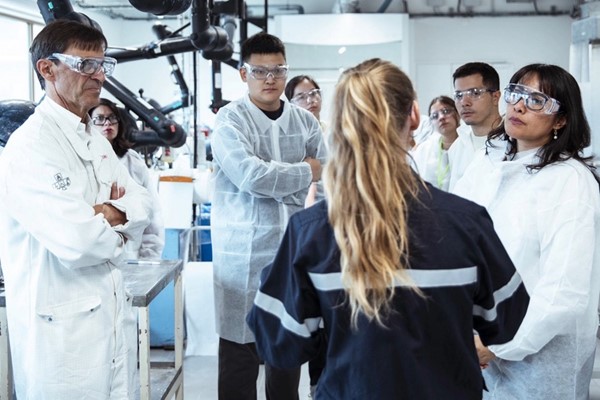 Several people in white lab coats and protective glasses  standing in a group in a laboratory setting